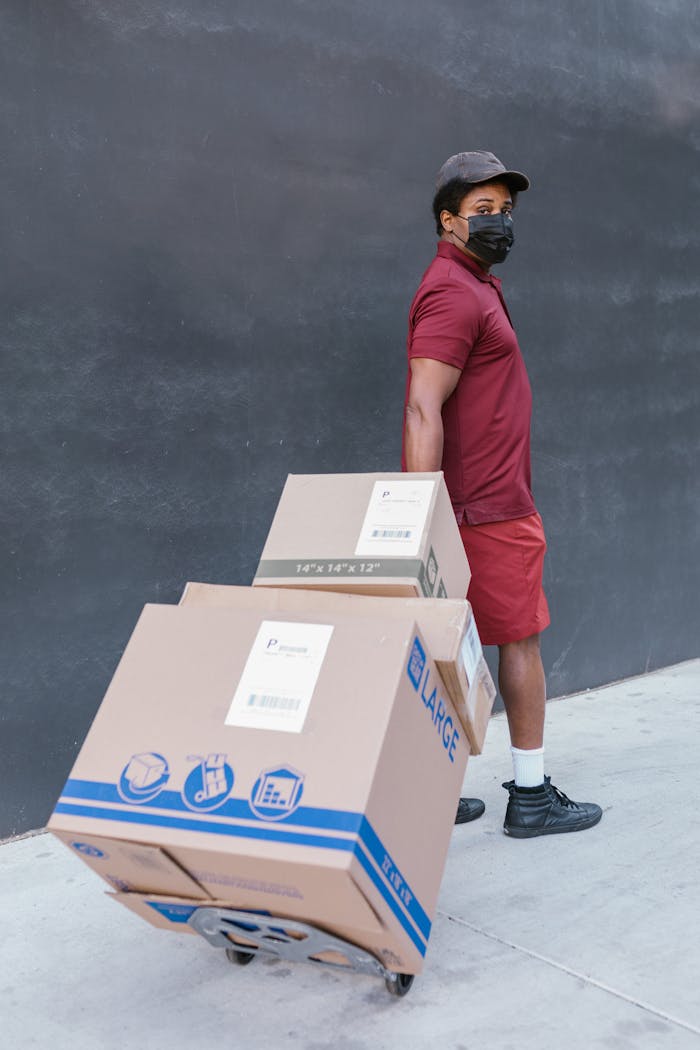gallery-04 An adult black man wearing a mask pulling a cart with large cardboard boxes outdoors.